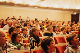 Erwachsene und viele Kinder in einem Theatersaal. Einige Kinder rufen etwas mit weit offenem Mund.